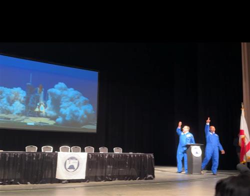 Dr. Nichols and Mr. Terrell in space flight suits by screen of a rocket launch